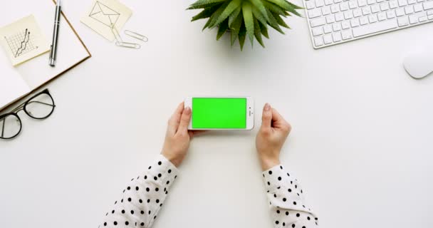 Vue de dessus sur le bureau blanc et smartphone blanc avec écran vert et mains féminines collant dessus. Horizontal. Des trucs de bureau à côté. Clé chromatique . — Video