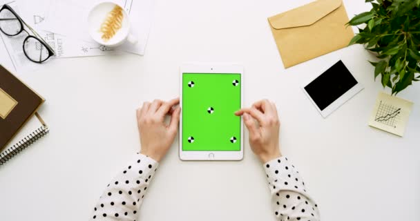 Ovanifrån på vita skrivbord och vit tablett dator med grön skärm och kvinnliga händer tejpa på den. Lodrät. Kontor grejer bredvid. Färgtransparens. Spåra rörelse — Stockvideo