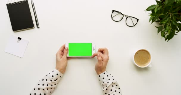 Вид зверху на офісний стіл і білий смартфон з зеленим екраном і жіночими руками, прикріпленими на ньому. Горизонтально. Офісні речі та кава поруч. Клавіша Chroma . — стокове відео
