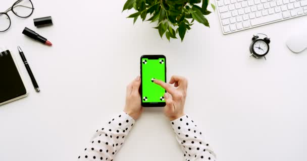 Vista dall'alto sulla scrivania bianca dell'ufficio e lo smartphone nero con schermo verde e le mani femminili che lo registrano. Verticale. Roba da ufficio. Chiave cromatica. Movimento di tracciamento — Video Stock