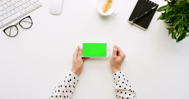 Vista superior na mesa do escritório branco e smartphone branco com tela verde e mãos femininas gravando nele. Horizontal. Coisas de escritório ao lado. Chave Chroma . — Vídeo de Stock