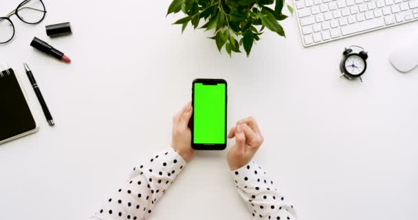 Felülnézet, fehér íróasztal és fekete smartphone-val a zöld képernyő és a női kéz szalagra rajta. Függőleges. Irodai cuccot mellett. Chroma-kulcs. — Stock videók