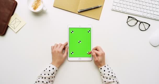 Vista superior en el escritorio de la oficina blanca y tableta blanca con pantalla verde y manos femeninas pegadas en ella. Vertical. Cosas de oficina al lado. Clave de croma. Movimiento de seguimiento — Vídeo de stock
