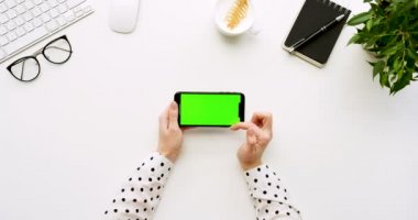 Üstten Görünüm beyaz Ofis Masası ve yeşil ekran ve üzerinde taping eller ile siyah smartphone. Yatay. Yanında başı işleri. Chroma anahtar.