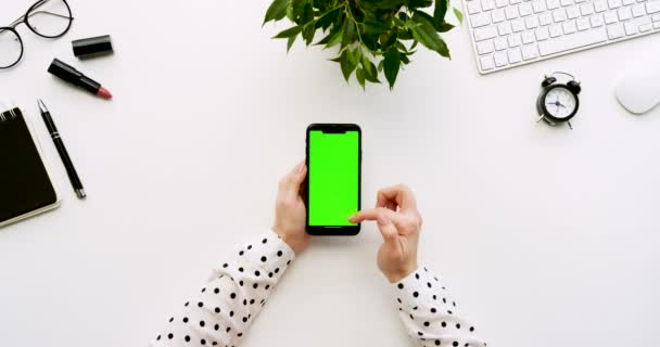 Ovanifrån på vita skrivbord och svart smartphone med grön skärm och kvinnliga händer tejpa på den. Lodrät. Kontor grejer bredvid. Färgtransparens. — Stockvideo