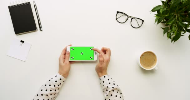 Top view on the office desk and white smartphone with green screen and female hands taping on it. Horizontal. Office stuff and coffee beside. Chroma key. Tracking motion — Stock Video