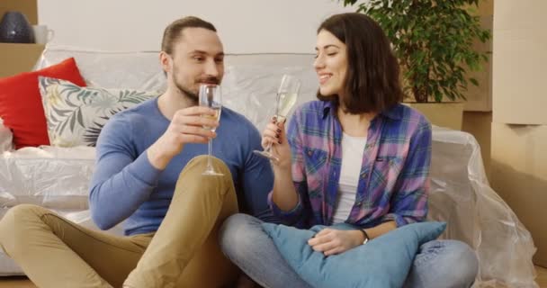 Junges glückliches frisch verheiratetes Paar, das auf dem Boden zwischen Logen im gemütlichen Wohnzimmer sitzt und Champagner trinkt, als es den Einzug in die neue Wohnung feiert. drinnen — Stockvideo