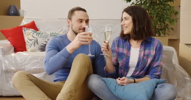 Young glimlachte vrouw en man zittend op de vloer omringd door verpakt vakken en thuis spullen in de kamer licht en drinken champagne als ze bewegen in de nieuwe flat. Binnenkant — Stockvideo