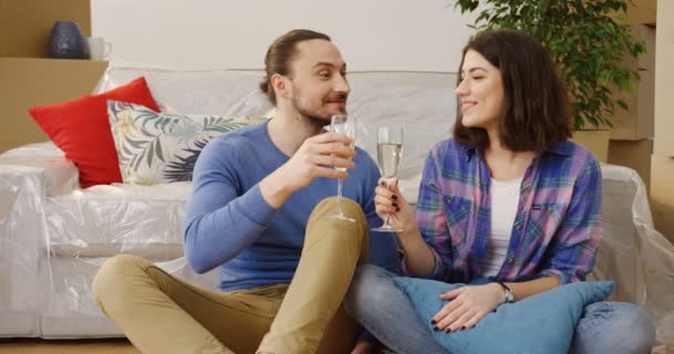 Молодой веселый только что женатый мужчина и женщина отдыхают на полу между коробками и домашними вещами в гостиной, обнимаются, улыбаются и пьют шампанское, празднуя вместе переезд в новый дом — стоковое видео
