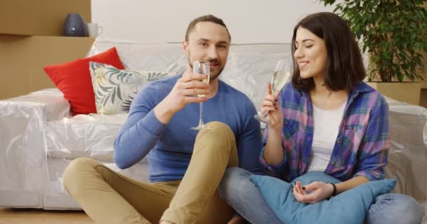 Joven atractiva mujer caucásica y hombre celebrando su mudanza al nuevo piso y beber champán mientras están sentados en el suelo entre cosas nuevas y cajas llenas. En interiores — Vídeos de Stock