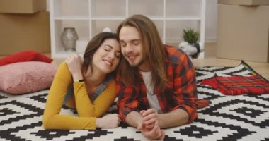 Genç ve güzel bir çift halının üzerinde uzanmış dinleniyor, yeni daireye taşınırken paketlenmemiş kutuların arasında gülüyor ve sohbet ediyorlar. Kapatın. Portre. İçeride.