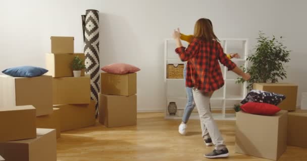 Joven pareja alegre caucásica bailando y peleando con almohadas entre cajas con sus cosas mientras se mueven en el nuevo piso moderno. De interior — Vídeo de stock