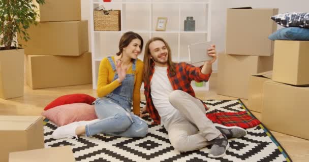 Young lovely couple sitting on the carpet on the floor among unpacked boxes, having videochat and showing their new apartment. Indoors — Stock Video