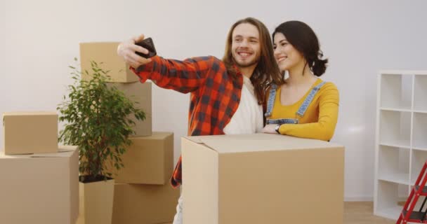 Молодая прекрасная пара позирует в уютной гостиной в окружении распакованных коробок, переезжая вместе и делая селфи на черном смартфоне. В помещении — стоковое видео