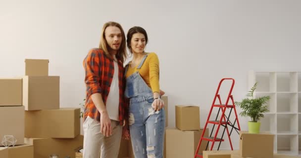 Молодая кавказская пара влюблена, стоя посреди милой комнаты, полной распакованных коробок, планируя дизайн квартиры, обнимаясь и улыбаясь в камеру. В помещении — стоковое видео