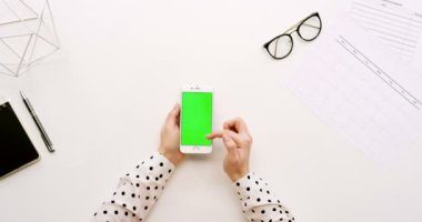 Üstten Görünüm Ofis Masası ve beyaz Smartphone ile yeşil perde ve eller üzerinde kayıt. Dikey. Yanında başı işleri. Chroma anahtar.