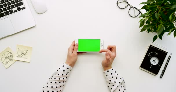 Top view on the office desk and white smartphone with green screen and female hands taping on it. Horizontal. Office stuff beside. Chroma key. — Stock Video