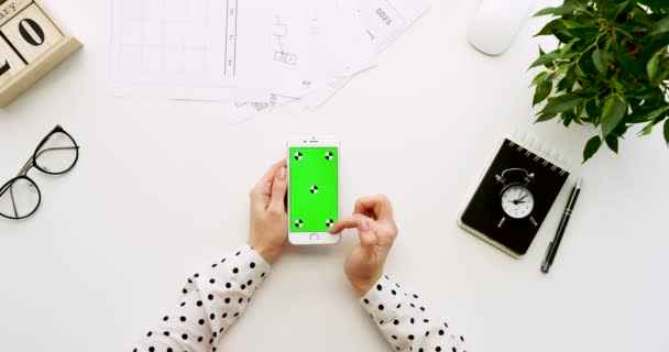 Ovanifrån på office skrivbord och vit smartphone med grön skärm och kvinnliga händer tejpa på den. Lodrät. Kontor grejer bredvid. Färgtransparens. Spåra rörelse — Stockvideo