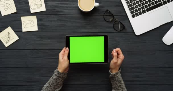 Vue de dessus sur le bureau noir et la tablette noire avec écran vert et mains féminines collant dessus. Horizontal. Des trucs de bureau, un ordinateur portable et un café à côté. Clé chromatique .