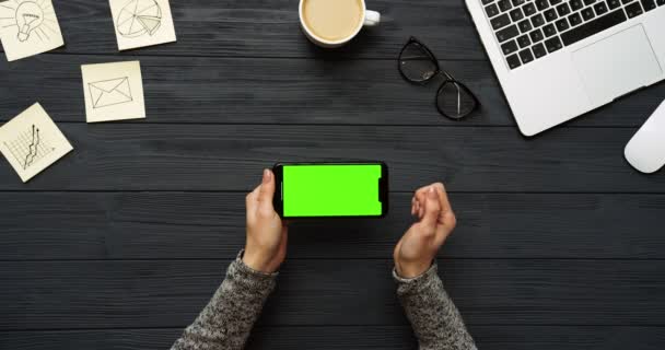 Vista dall'alto sulla scrivania dell'ufficio nero e lo smartphone nero con schermo verde e le mani femminili che lo registrano. Orizzontale. Roba da ufficio, laptop e caffe 'accanto. Chiave cromatica . — Video Stock