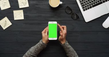 Üstten Görünüm siyah Ofis Masası ve beyaz Smartphone ile yeşil perde ve eller üzerinde kayıt. Dikey. Başı işleri, dizüstü bilgisayar ve kahve yanında. Chroma anahtar.