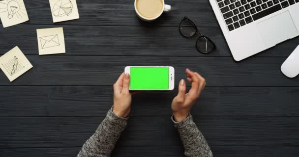 Visão superior na mesa do escritório preto e smartphone branco com tela verde e mãos femininas gravando sobre ele. Horizontal. Material de escritório, laptop e café ao lado. Chave Chroma . — Vídeo de Stock