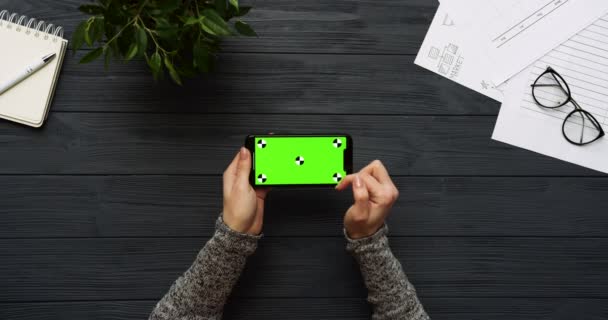 Ovanifrån på svart skrivbord och svart smartphone med grön skärm och kvinnliga händer tejpa på den. Horisontella. Kontor grejer bredvid. Färgtransparens. Spåra rörelse — Stockvideo