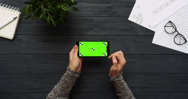 Ovanifrån på svart skrivbord och svart smartphone med grön skärm och kvinnliga händer tejpa på den. Horisontella. Kontor grejer bredvid. Färgtransparens. Spåra rörelse — Stockvideo