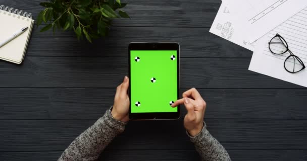 Vista superior en el escritorio negro de la oficina y la tableta negra con la pantalla verde y las manos femeninas pegadas en ella. Vertical. Cosas de oficina al lado. Clave de croma. Movimiento de seguimiento — Vídeo de stock