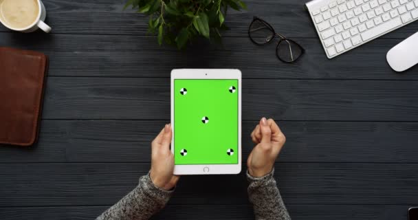 Vista superior en el escritorio de oficina negro y tableta blanca con pantalla verde y manos femeninas pegadas en ella. Vertical. Cosas de oficina al lado. Clave de croma. Movimiento de seguimiento — Vídeo de stock