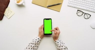 Üstten Görünüm beyaz Ofis Masası ve yeşil ekran ve üzerinde taping eller ile siyah smartphone. Dikey. Yanında başı işleri. Chroma anahtar.