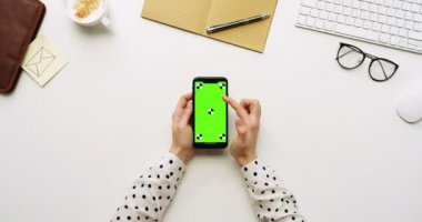 Üstten Görünüm beyaz Ofis Masası ve yeşil ekran ve üzerinde taping eller ile siyah smartphone. Dikey. Yanında başı işleri. Chroma anahtar. Hareket izleme