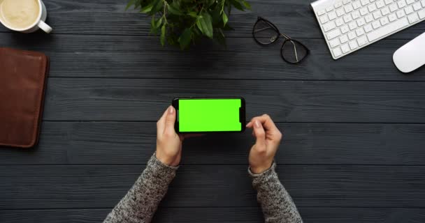 Vista superior en el escritorio negro de la oficina y el teléfono inteligente negro con pantalla verde y manos femeninas pegadas en él. Horizontal. Cosas de oficina al lado. Clave de croma . — Vídeos de Stock