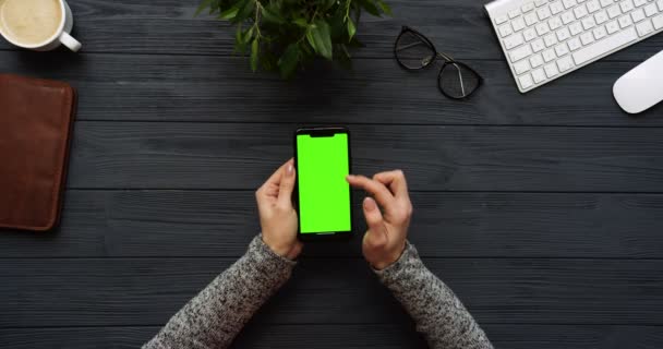 Ovanifrån på svart skrivbord och svart smartphone med grön skärm och kvinnliga händer tejpa på den. Lodrät. Kontor grejer bredvid. Färgtransparens. — Stockvideo