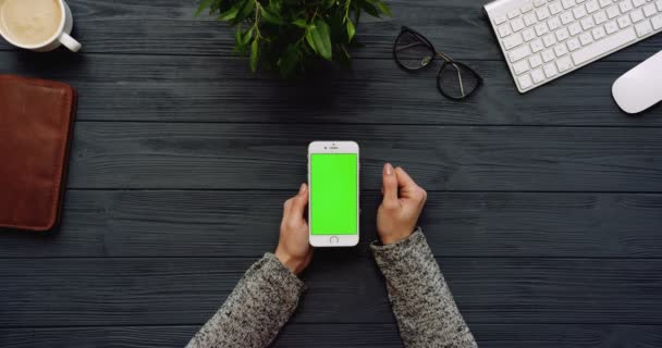 Ovanifrån på svart office skrivbord och vit smartphone med grön skärm och kvinnliga händer tejpa på den. Lodrät. Kontor grejer bredvid. Färgtransparens. — Stockvideo