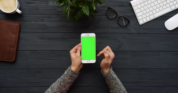 Vista superior en el escritorio negro de la oficina y el teléfono inteligente blanco con pantalla verde y las manos femeninas pegadas en él. Vertical. Cosas de oficina al lado. Clave de croma . — Vídeo de stock