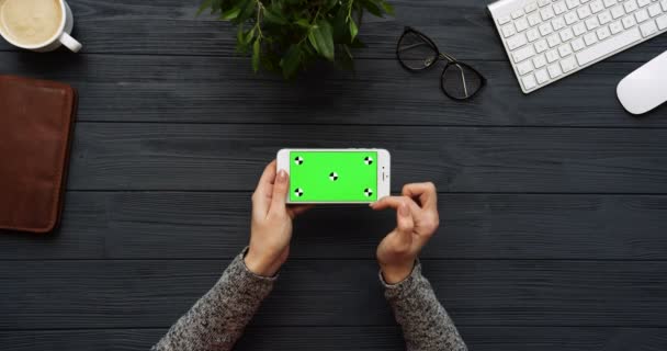 Top view on the black office desk and white smartphone with green screen and female hands taping on it. Horizontal. Office stuff beside. Chroma key. Tracking motion — Stock Video