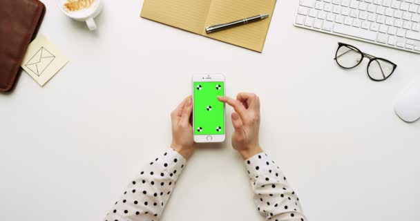 Top view on the white office desk and white smartphone with green screen and female hands taping on it. Vertical. Office stuff beside. Chroma key. Tracking motion — Stock Video