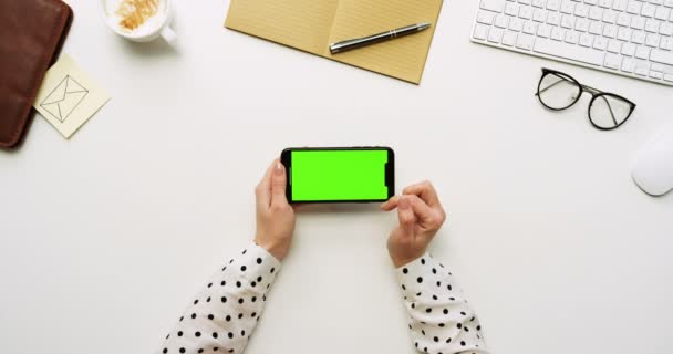 Top view on the white office desk and black smartphone with green screen and female hands taping on it. Horizontal. Office stuff beside. Chroma key. — Stock Video