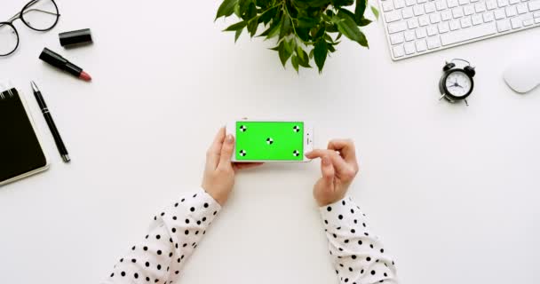 Top view on the white office desk and white smartphone with green screen and female hands taping on it. Horizontal. Office stuff beside. Chroma key. Tracking motion — Stock Video