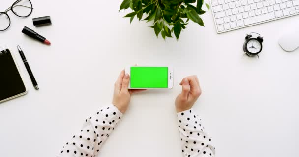 Top view on the white office desk and white smartphone with green screen and female hands taping on it. Horizontal. Office stuff beside. Chroma key. — Stock Video