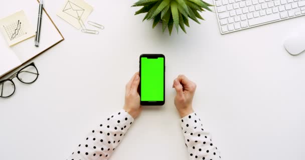 Вид зверху на білий офісний стіл і чорний смартфон з зеленим екраном і жіночими руками, прикріпленими на ньому. Вертикально. Офісні речі поруч. Клавіша Chroma . — стокове відео