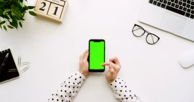 Üstten Görünüm beyaz Ofis Masası ve yeşil ekran ve üzerinde taping eller ile siyah smartphone. Dikey. Başı işleri, dizüstü bilgisayar ve gözlük yanında. Chroma anahtar.