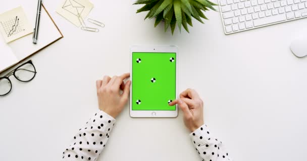Ovanifrån på vita skrivbord och vit tablett dator med grön skärm och kvinnliga händer tejpa på den. Lodrät. Kontor grejer bredvid. Färgtransparens. Spåra rörelse — Stockvideo