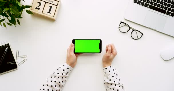 흰색 사무실 책상 및 녹색 스크린 및 여성의 손에 그것에 녹화 블랙 스마트폰 최고 볼 수 있습니다. 수평. 오피스 물건, 노트북와 옆에 안경입니다. 크로마 키. — 비디오