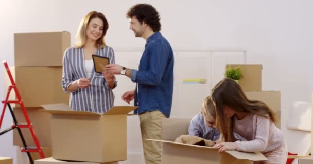 Jovens pais felizes com seus filhos conversando enquanto desempacotam caixas com coisas de casa em um movimento no dia. No novo apartamento. Interior — Vídeo de Stock
