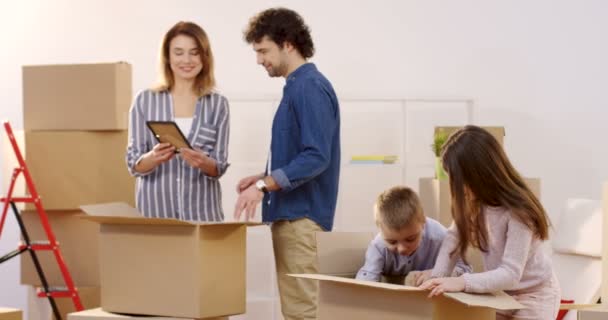 Gelukkige familie van ouders en kinderen vakken met home spullen uitpakken tijdens het verplaatsen in het nieuwe appartement. Indoor — Stockvideo