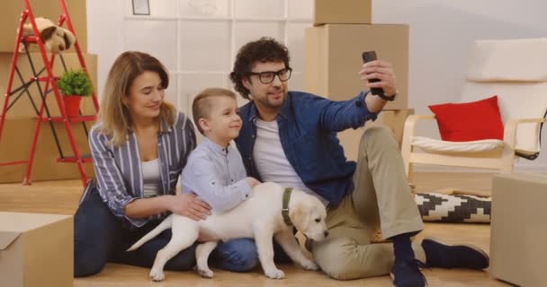 Happy Caucasian family sitting on the floor with a little labrador puppy and doing selfies surrounded by unpacked boxes in their new home. Indoors — Stock Video