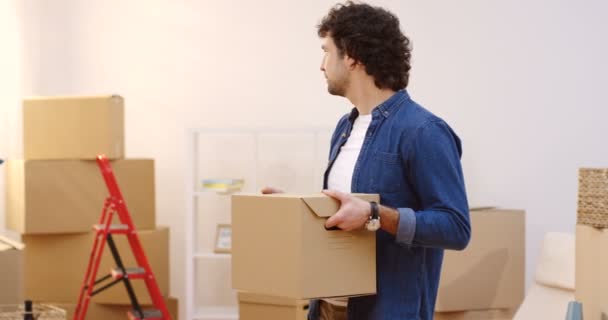 Portrait of the attractive manin a jeans shirt smiling and looking to the camera with a carton box in hands in the new apartment on a moving in day. Inside — Stock Video