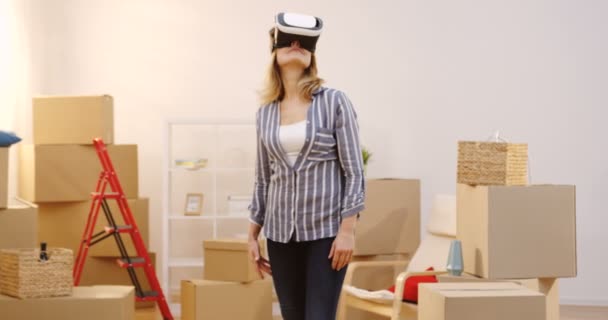 Mulher bonita em óculos VR com um fone de ouvido VR na sala de estar cheia de caixas de uma nova casa durante a mudança. Interior — Vídeo de Stock